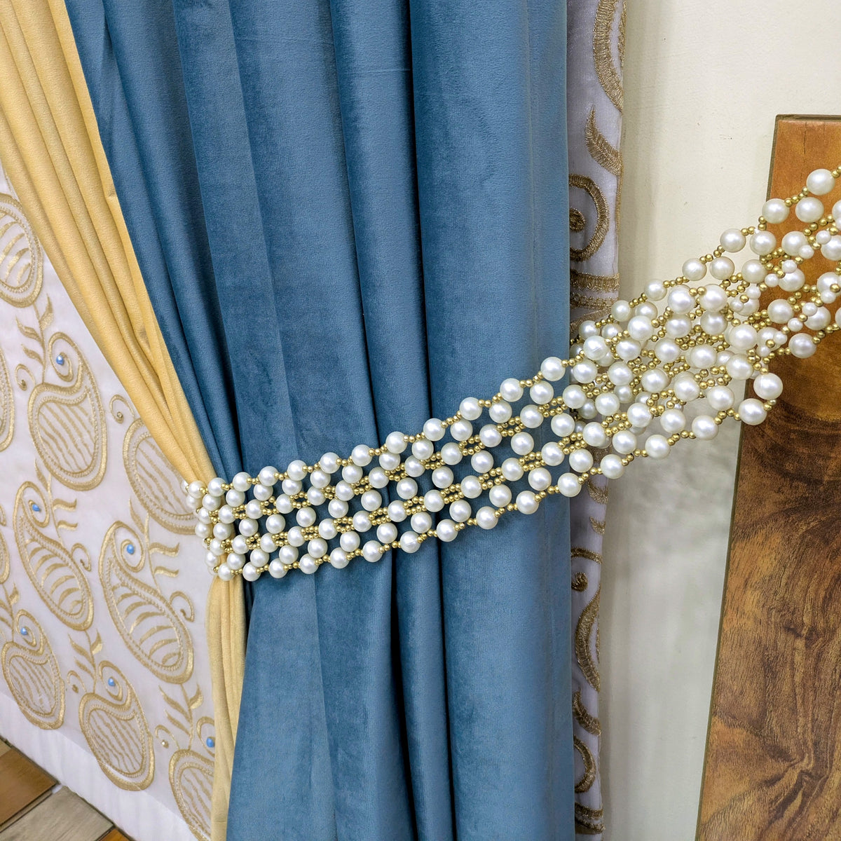 A close-up of a blue velvet curtain tied back with a decorative pearl and gold tieback band, with patterned drapes and a wooden panel in the background.