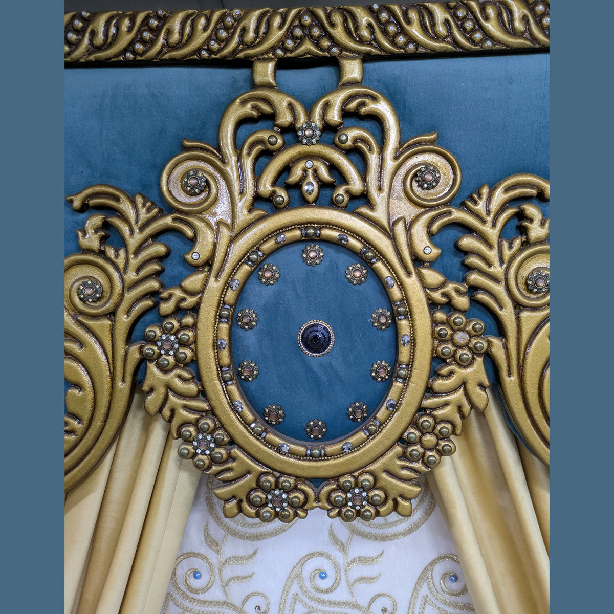 Close-up of an ornate upholstered headboard or decorative furniture panel with a blue velvet center, gold carved scrollwork, jeweled details, and draped gold fabric below.