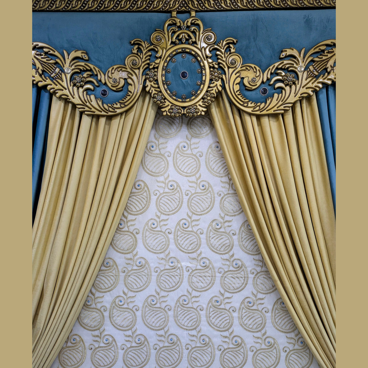 An ornate window curtain valance set featuring gold drapes with a central decorative gold medallion, flanked by blue side panels and a blue top band, set against a white patterned inner curtain.