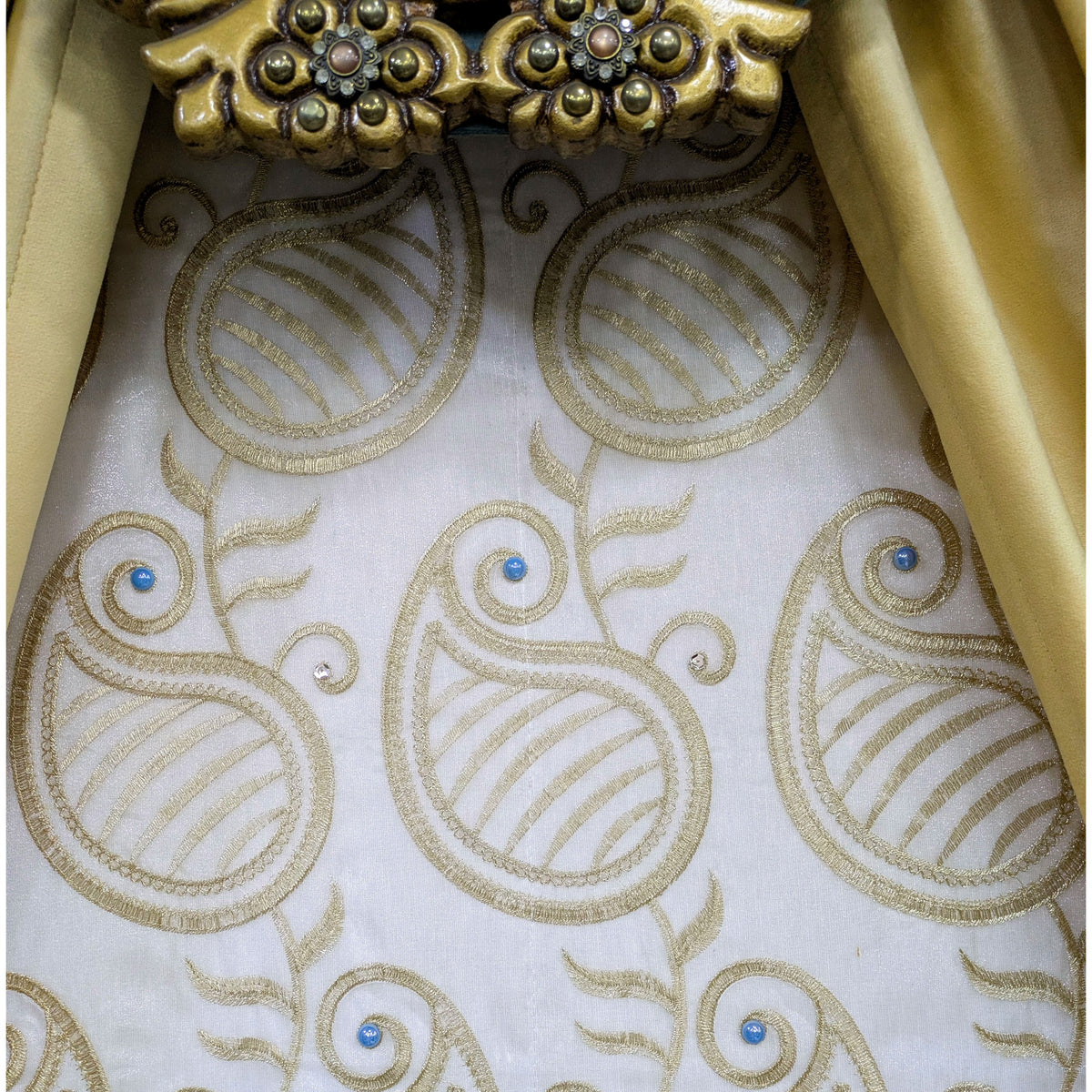 Close-up of a white fabric with gold paisley embroidery and small blue bead accents, framed by gold drapery and an ornate gold decorative element at the top.