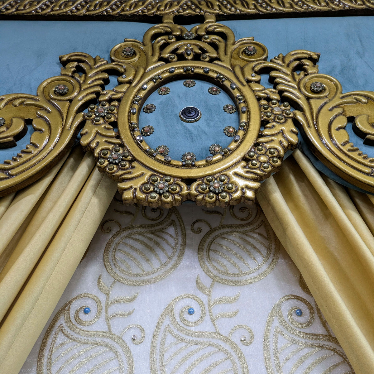 An ornate gold decorative headboard frame with carved scrollwork and a central oval medallion, flanked by draped cream satin curtains over a patterned cream and gold upholstered headboard surface.
